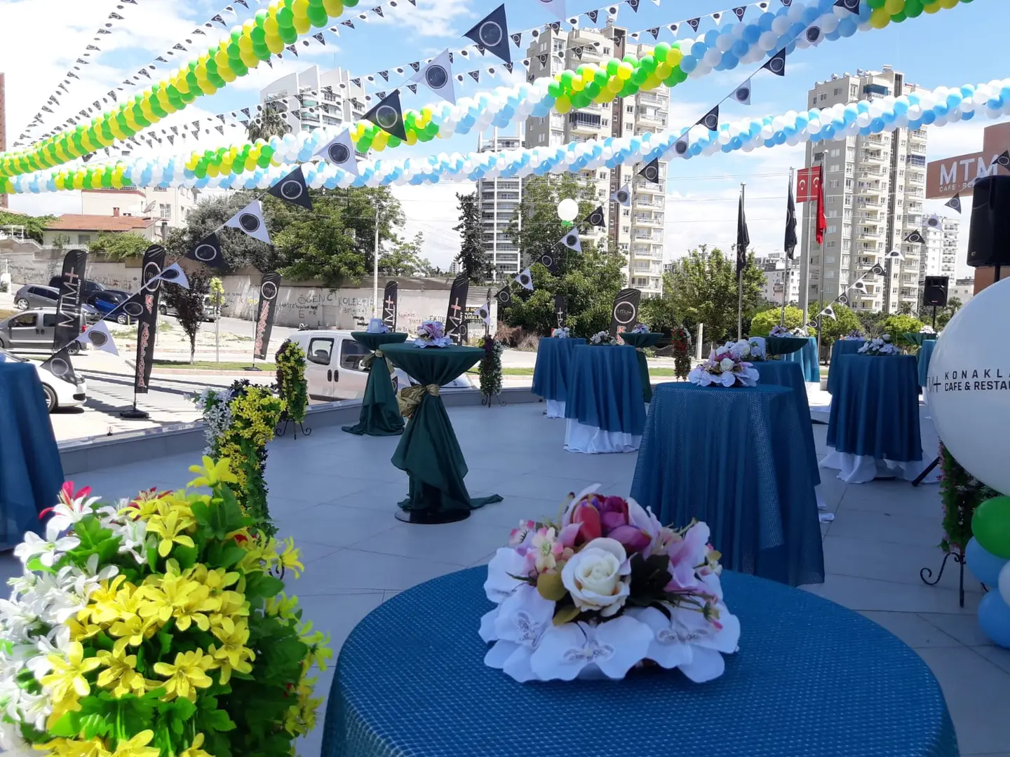 Açık alanda kurulan kokteyl düzeni: örtülü bistro masalar, çiçek aranjmanı ve flama süsleme.